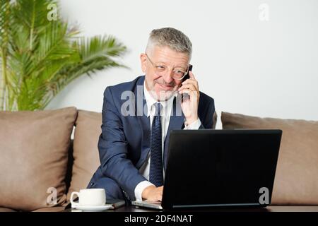 Portrait of smiling senior businessman talking on phone and reading e-mail on laptp screen Stock Photo