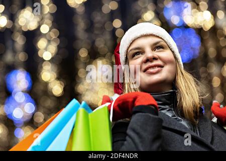 A woman with long hair and a Santa hat near the window of a city store with purchases in colorful, paper bags. New year's shopping. Stock Photo