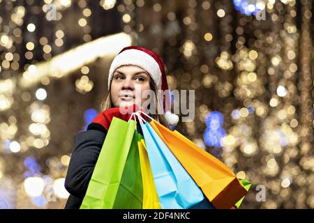 A woman with long hair and a Santa hat near the window of a city store with purchases in colorful, paper bags. New year's shopping. Stock Photo