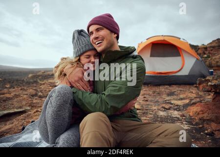 Happy young man hugging his smiling girlfriend and looking away during camping Stock Photo