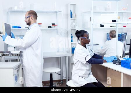 African scientist working on computer wearing protection gear doing analysis. Multiethnic team of researchers working in microbiology lab testing solution for medical purpose. Stock Photo