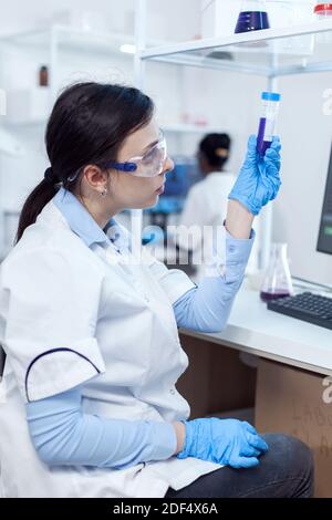 Young scientist analysing scientific sample on plastic test tube with african coworker sitting at her workplace. Chemistry researcher in sterile laboratory using modern technology to test microbiology liquid. Stock Photo