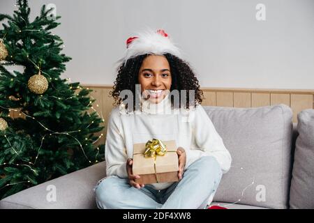 Attractive millennial african woman in santa hat staying at home and smiling, holding gift box with gold ribbon. Celebrate Christmas and New Year at Stock Photo