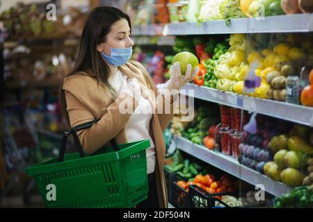 Happy woman in protective mask taking fresh vegetables while standing by groceries in supermarket. Beautiful young girl with food basket choosing food Stock Photo