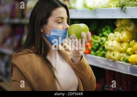 Happy woman in protective mask taking fresh vegetables while standing by groceries in supermarket. Beautiful young girl with food basket choosing food Stock Photo