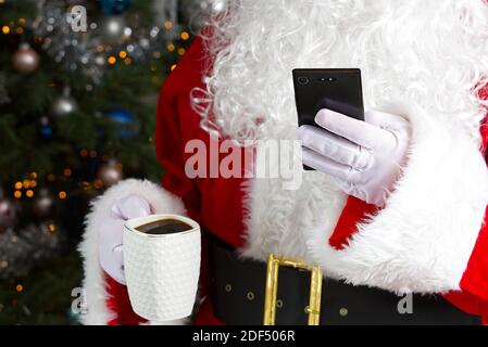 Santa claus holding white mug with coffee and smartphone in his hand, close-up. mobile Phone And Coffee mug in santa hands. Stock Photo