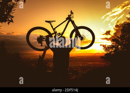 Silhouette of a mountain biker holding a bicycle over his head at sunset. MTB, enduro, freeride background. Stock Photo