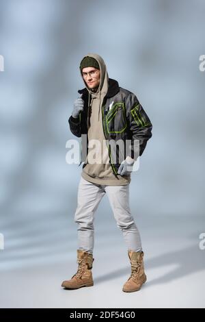 full length of young trendy man in hat, hoodie and anorak standing on grey Stock Photo