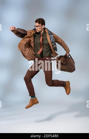 stylish man in winter coat holding leather briefcase while jumping on grey Stock Photo