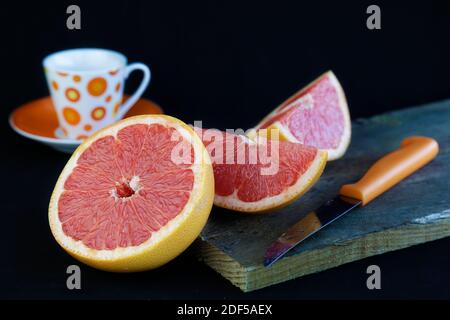 Grapefruit on a wooden cutting board, next to a knife and a cup of coffee against black background Stock Photo