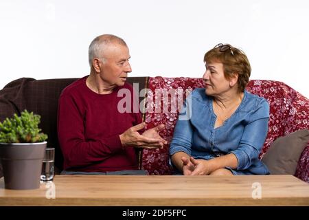 The old couple are sitting on couch, talking. The man gesturing with hands. Stock Photo