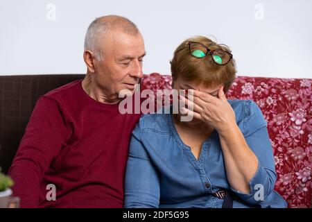 Elderly spouses are sitting on the couch, husband consoles disappointed wife. Stock Photo