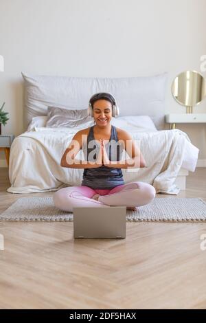 African American woman meditates at home in front of a laptop monitor. She listens to relaxing music through headphones. Vertical photo. Stock Photo