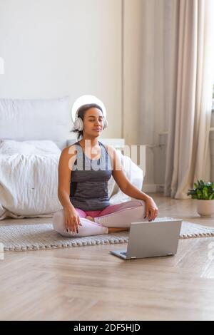 Vertical photo of an African-american woman meditates at home in front of a laptop monitor and listening to the music via headphones. Stock Photo