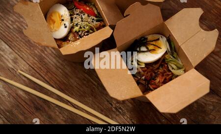 Chinese noodles with chicken and vegetables in cardboard boxes on a wooden background, Asian food delivery, concept of street food, copy space Stock Photo