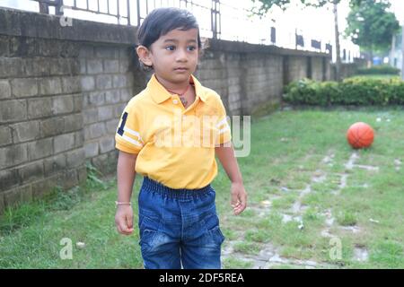 Indian cute little cheerful brunette Tamil baby boy in vibrant yellow casual wear playing with himself with an orange ball in green natural background Stock Photo