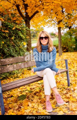 Full length shot of middle aged woman wearing casual clothes while sitting in bench and relaxing in the park. Fall weather. Stock Photo