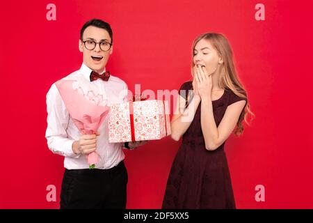 A handsome student, wearing glasses, holding books in her hands, gives a gift and a bouquet of flowers to his girlfriend against a red background. Val Stock Photo