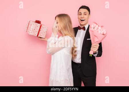 Handsome man in a suit making a surprise to a woman, closing her eyes with her hands on a pink background Stock Photo