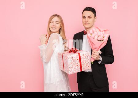 Elegant man in a suit makes a surprise to a woman, gives a bouquet of flowers and a box with a gift, on a pink background, concept of women's day Stock Photo