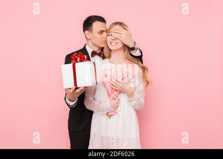 Handsome man in a suit making a surprise to a woman, closing her eyes with her hands on a pink background Stock Photo