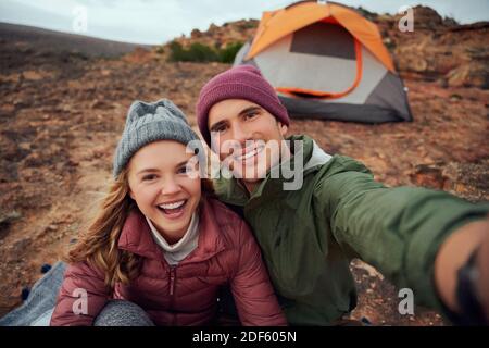 Cheerful young man and woman in winter clothes embracing and laughing while taking selfie during camping Stock Photo