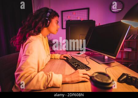 Side view of african american gamer in headset playing video game on computer with blank screen near joystick and coffee to go on blurred foreground Stock Photo