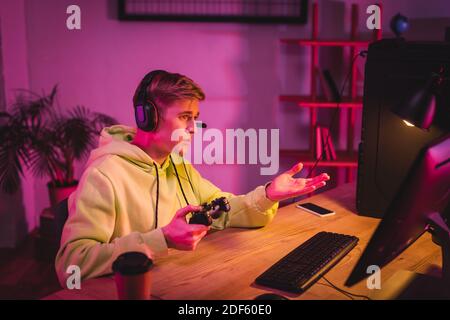 KYIV, UKRAINE - AUGUST 21, 2020: Gamer in headset holding joystick while pointing at computer monitor near smartphone and coffee to go on blurred fore Stock Photo