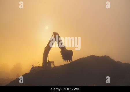 An excavator performs work in rubble in hazy and dusty early morning conditions. Stock Photo