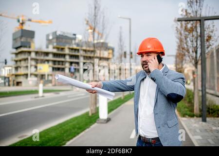 Young architect holding mobile phone and blueprint while looking away. Stock Photo