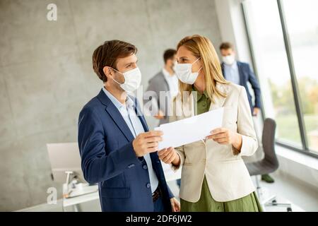 Young man and woman with protective facial masks discussing with paper in hands indoors in the office with young people works behind them Stock Photo