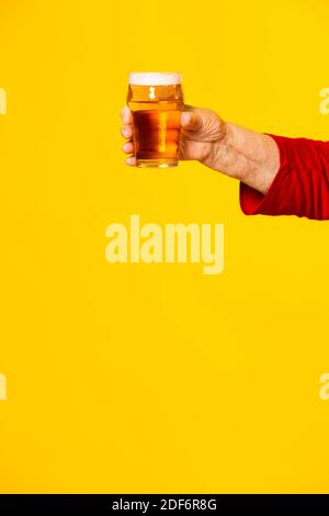 Hand of a senior woman holding a glass of half a pint of beer, against a yellow background Stock Photo