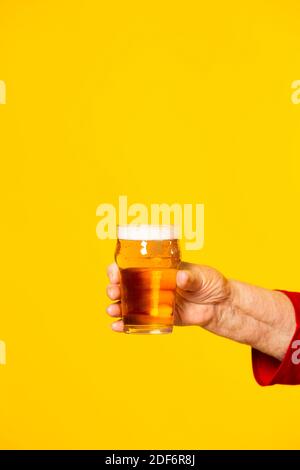 Hand of a senior woman holding a glass of half a pint of beer, against a yellow background Stock Photo