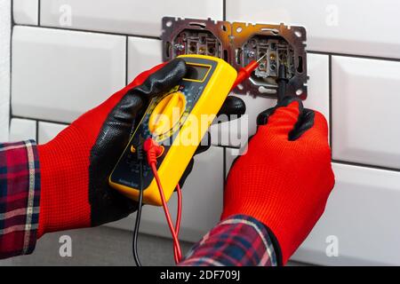 A guy in red work gloves, using a multimeter - tester, measures the voltage in the electrical network. Electrical outlet repair. Electrical outlet Stock Photo