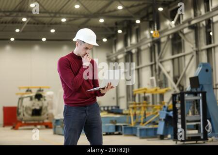 Portrait of a male factory manager in a white hard hat and red sweater holding laptop and mobile phone. Controlling the work process in the helicopter manufacturer. . High quality photo Stock Photo