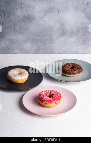 Three donuts on plates, chocolate, pink and vanilla donut with sprinkles, sweet glazed dessert food on white concrete textured background, angle view Stock Photo