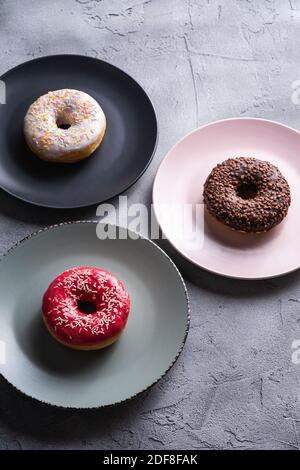 Three donuts on plates, chocolate, pink and vanilla donut with sprinkles, sweet glazed dessert food on concrete textured background, angle view Stock Photo
