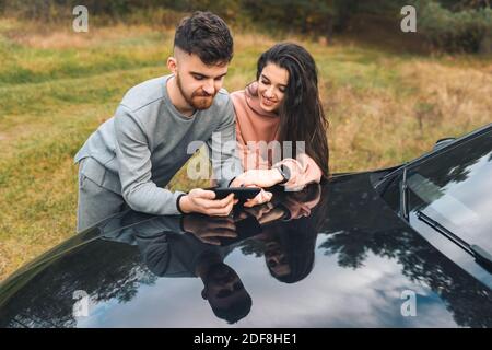 Young couple is looking at something cheerful in the smartphone, standing leaning on the hood of car Stock Photo