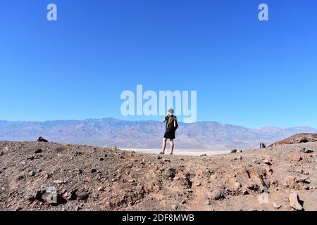 A male hiker wearing a backpack stands on a small hill at the trail head for Natural Bridge Canyon overlooking Badwater Basin and Death Valley, USA Stock Photo