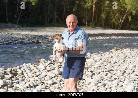 Senior man holding small wet Jack Russell terrier dog in his hand, after she swam in the near river on sunny day Stock Photo