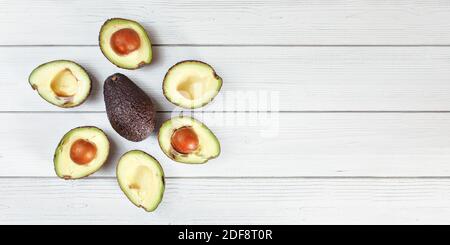 Ripe avocado halves arranged around whole dark brown pear (hass / bilse fruit variety) on white boards desk, banner with space for text right Stock Photo