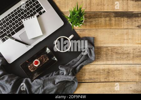 Laptop, small spiral notepad, butterscotch cake and cup of coffee on rustic wood. Flat lay. Stock Photo
