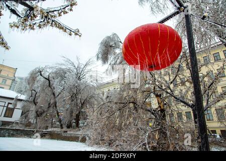 A Chinese red lantern hangs on the street in winter, it is very icy, icicles hang. Against the background of light bulbs in the city. Stock Photo