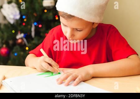 Cute child boy wearing red hat write letter to Santa sitting by desk, Christmas tree on background Stock Photo