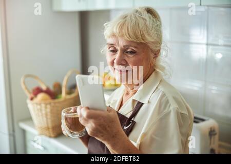 Nice old woman using cellphone and drinking tea at home Stock Photo