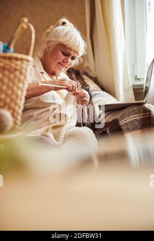 Nice old woman knitting and using notebook at home Stock Photo