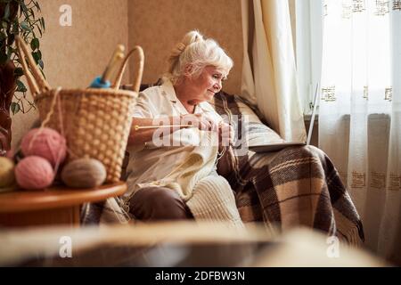 Lovely old woman knitting and using laptop at home Stock Photo