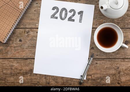 Year 2021 drawn on sheet of paper, upon rustic desk with teapot, tea cup, notepad and pencil, high angle view Stock Photo