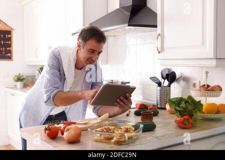Man cooking leaning on the kitchen bench full of vegetables consulting recipes on a tablet Stock Photo