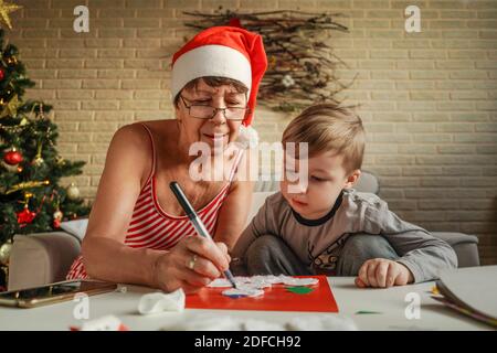 A Little boy with grandmother make Christmas craft, greeting card. Christmas tree and decoration on background. Stock Photo